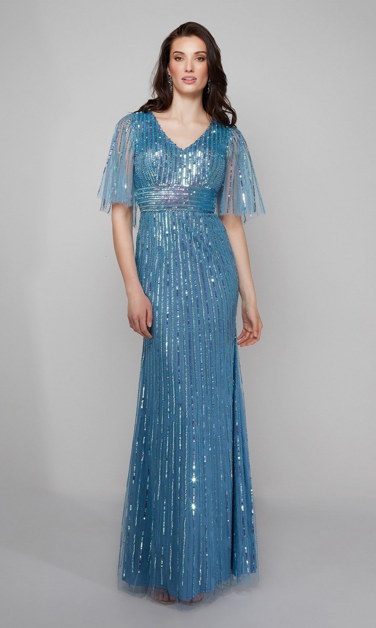 A woman poses against a plain gray background wearing the Alyce 27599 long formal dress, featuring blue beaded sequins, a floor-length silhouette, short sheer sleeves, and a V-neck.